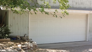 Eine Garage mit weißer Tür, ein Baum davor und einem Steinbruch.