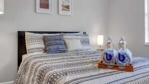 A neatly made bed with patterned bedding, two framed pictures on the wall, and a bedside table with water bottles.