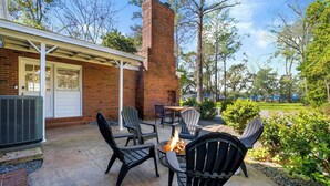 An outdoor patio with a fire pit and chairs, a brick building with a chimney, and a view of a grassy area.