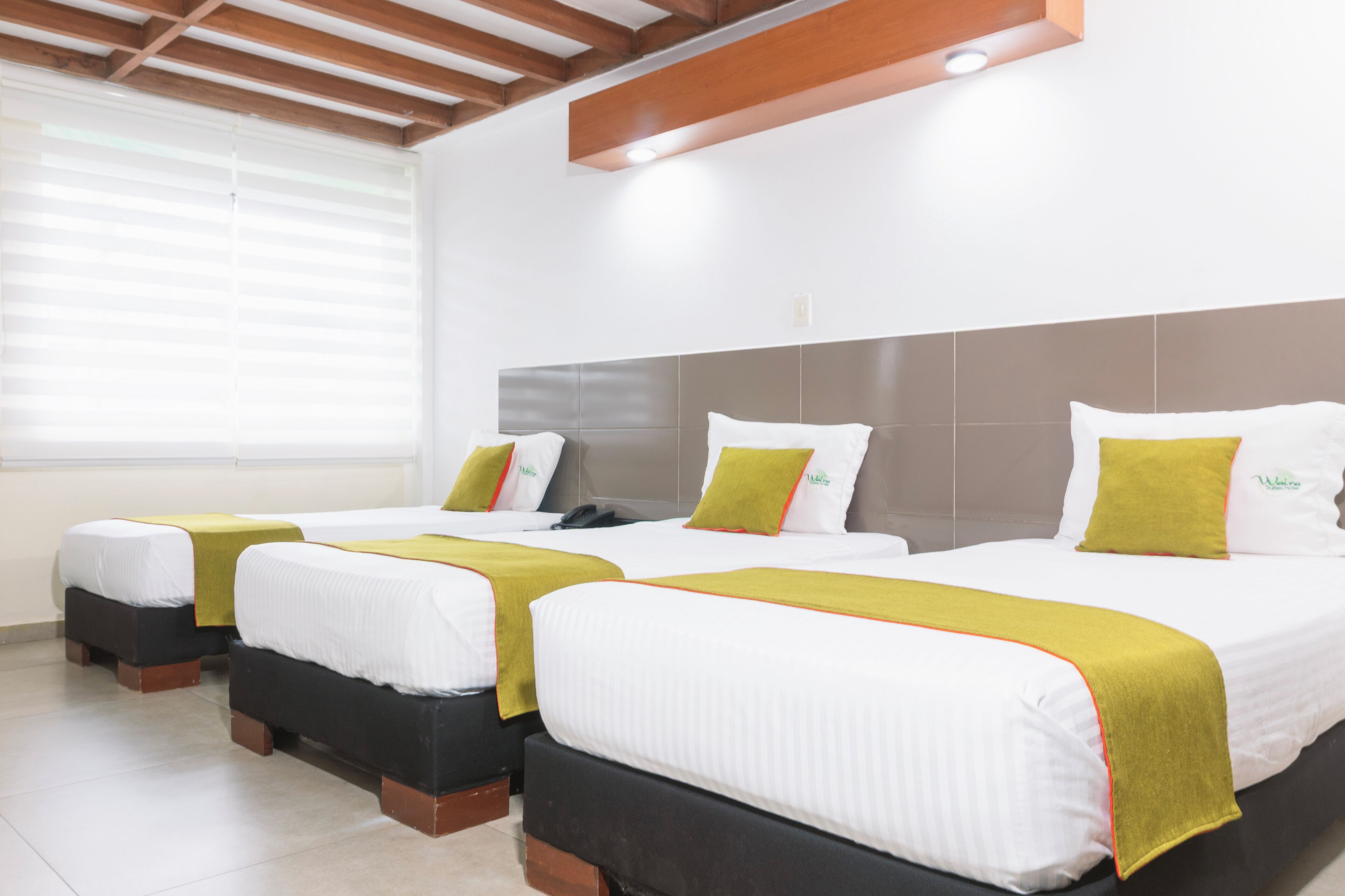 A hotel room with three beds, each with white bedding and green pillows. The room has a wooden ceiling and a large window with blinds.