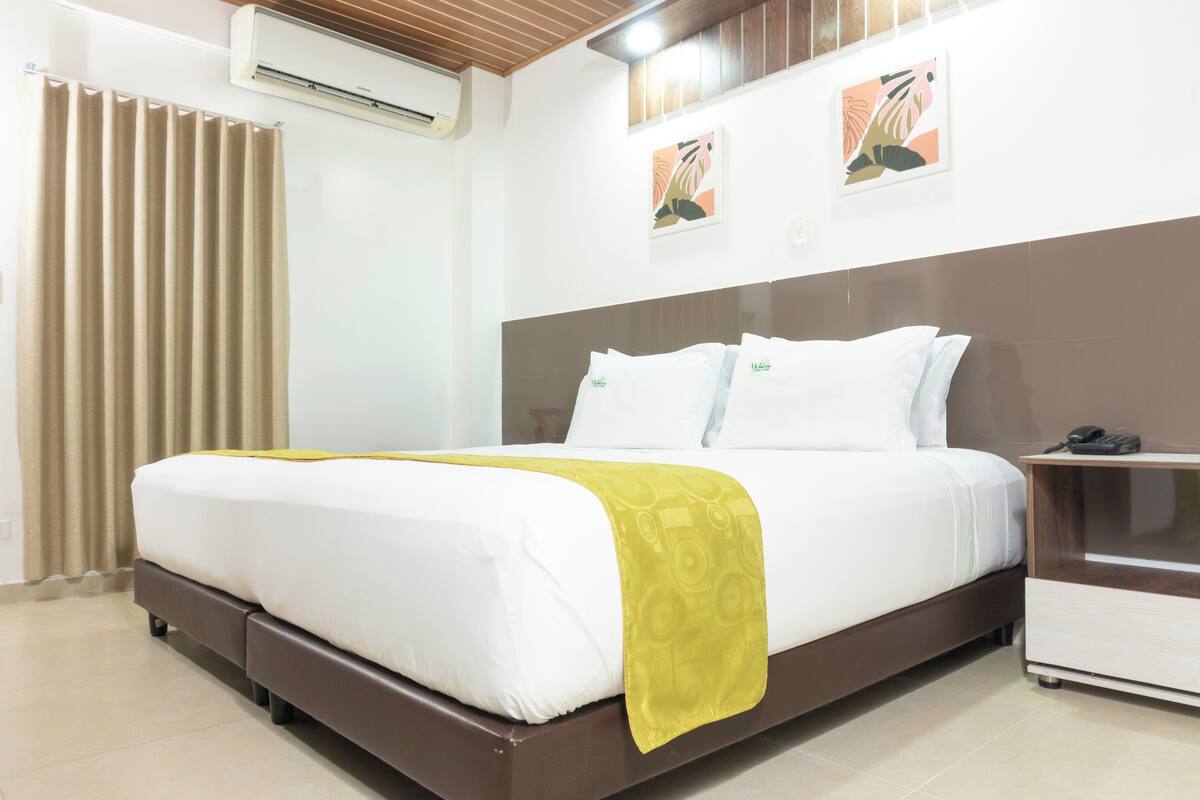 Una cama bien tendida con una colcha blanca y amarilla, un cabecero de madera y una mesita de noche con un teléfono.