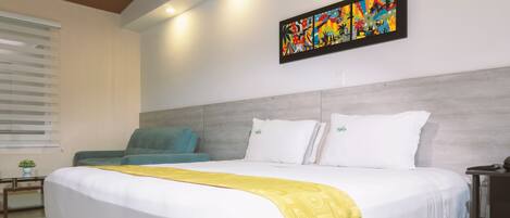 A hotel room with a large bed, a teal armchair, a colorful abstract painting on the wall, and a small plant on the nightstand.