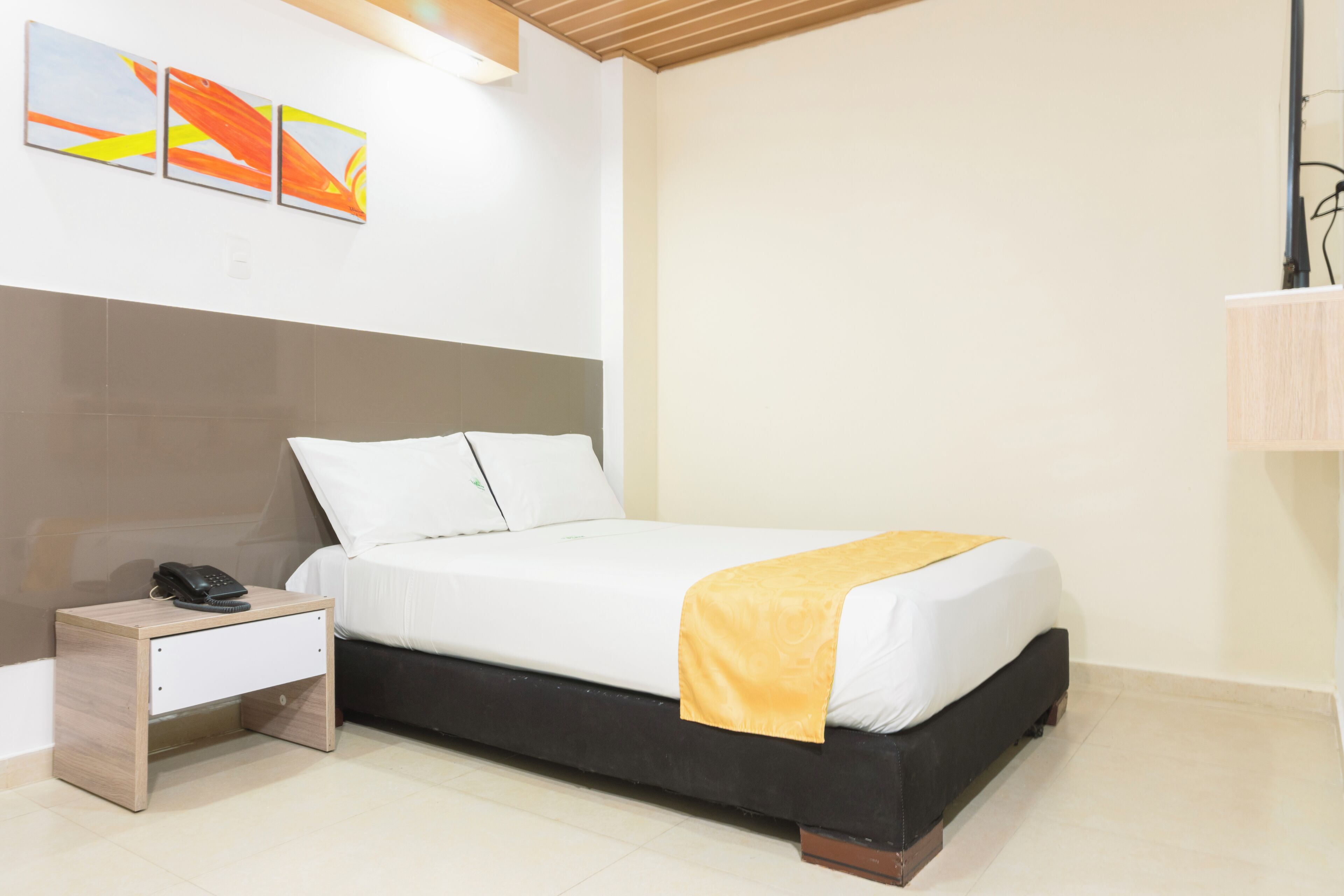 A neatly made bed with a white and yellow bedspread, a bedside table with a phone, and abstract wall art.