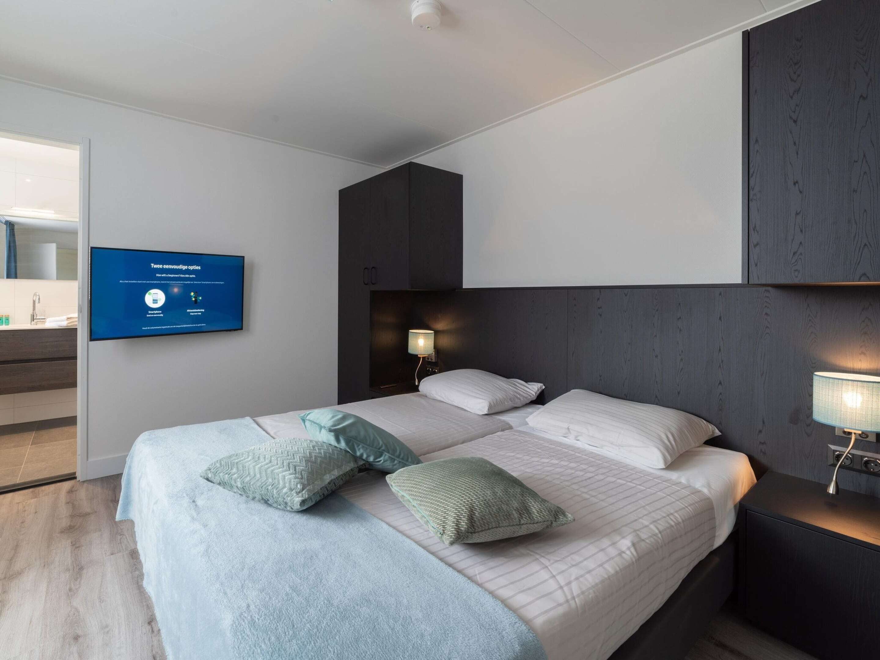 A modern hotel room with a large bed, two bedside tables, a mounted TV, and a dark wood headboard.
