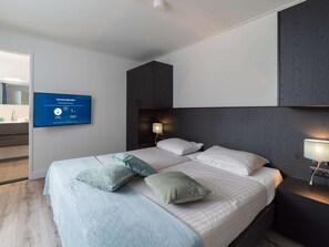 Quarto de hotel moderno com uma cama grande, duas mesinhas de cabeceira, uma TV na parede e uma cabeceira de madeira escura.