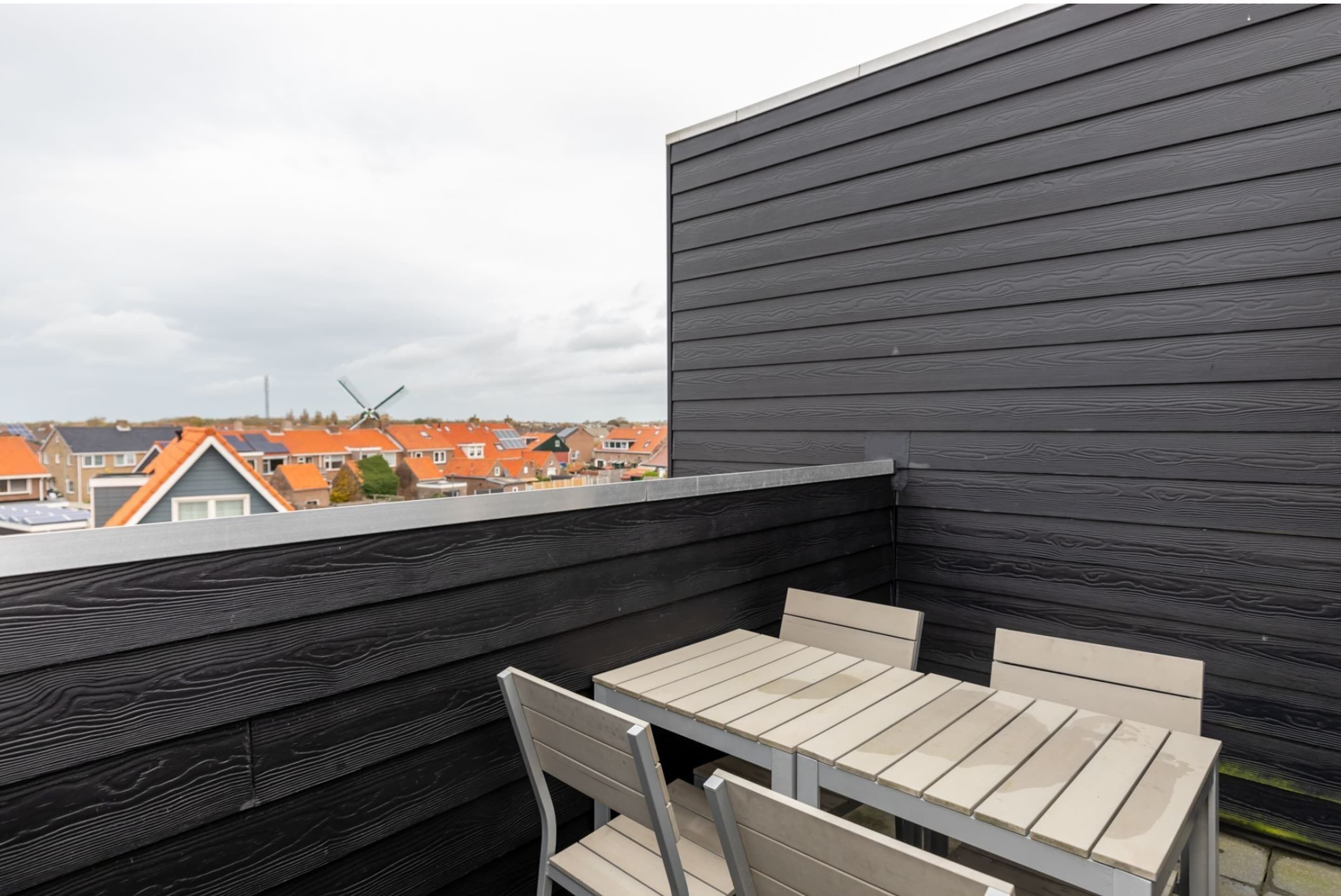 Une terrasse sur le toit, avec une table et des chaises, d’où l’on peut admirer un quartier résidentiel avec des maisons aux toits orange et une éolienne.