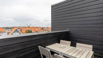 A rooftop terrace with a table and chairs, overlooking a residential area with orange-roofed houses and a wind turbine.