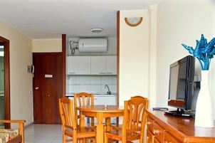 Egy étkezősarok, melyben egy fa asztal és székek találhatók, egy televízió egy fa állványon, és a háttérben egy konyha látható.