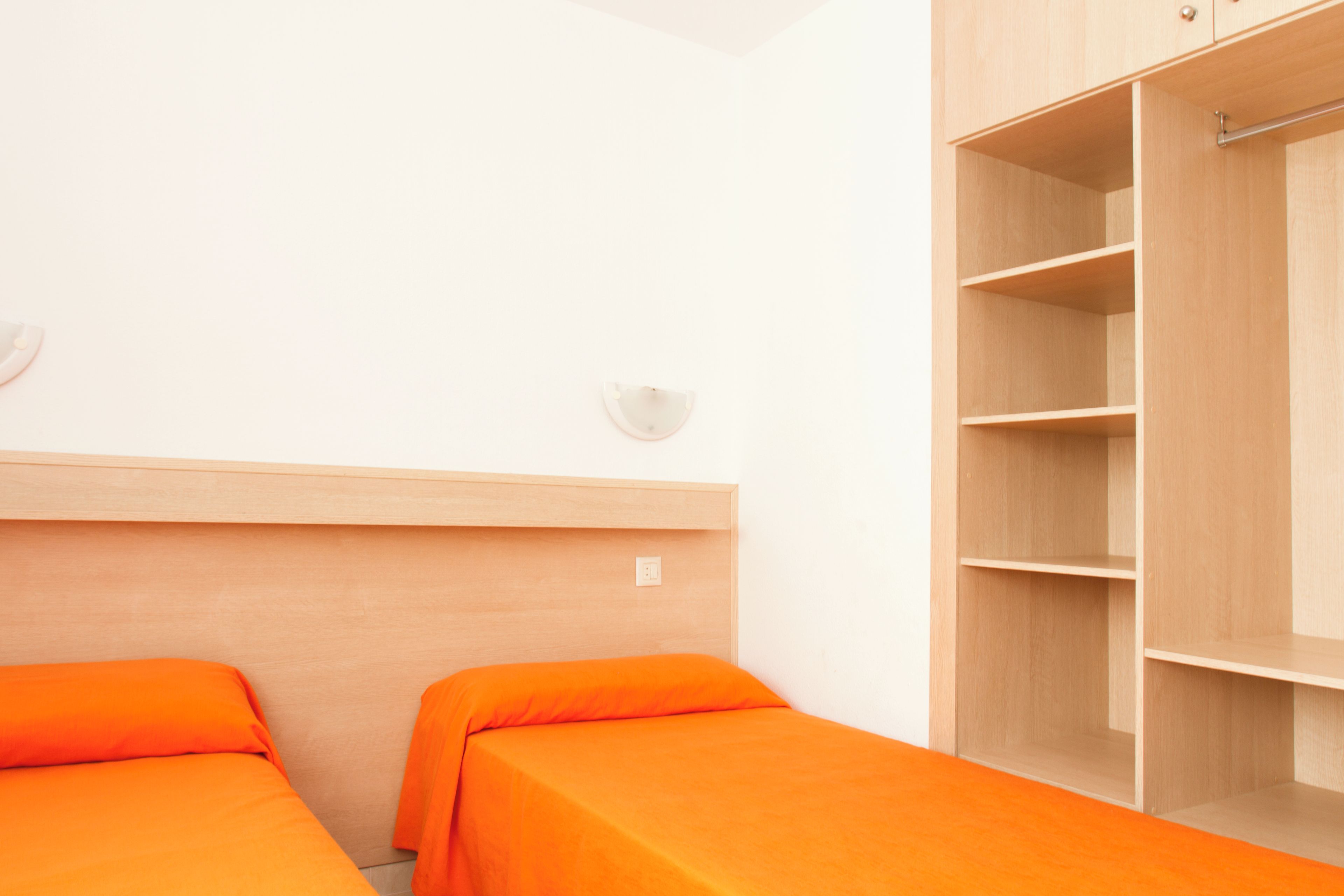 Una habitación con dos camas, cada una con colcha naranja, cabecero de madera y un armario con repisas abiertas.