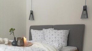 A bed with a wooden frame, white bedding, and geometric-patterned pillows. There is a small plant and a candle on the bedside table. - Apartment 2- NEST'L Apartments (Mösern)