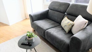 A grey sofa with two pillows, a small round table with a plant and a remote control, and a light-colored rug. - Apartment 2- NEST'L Apartments (Mösern)