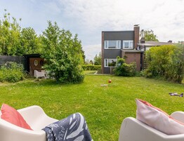 Un patio trasero residencial con una casa de dos pisos, un cobertizo pequeño y un césped con una silla y cojines blancos.