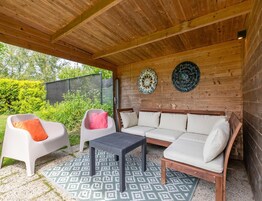 Un área de estar al aire libre con techo de madera, dos sillas blancas con cojines, una mesa de tejido gris y una alfombra estampada.