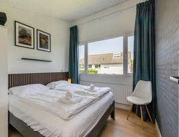 Habitación de hotel con una cama, una silla, un ventanal con cortinas y dos cuadros en la pared.