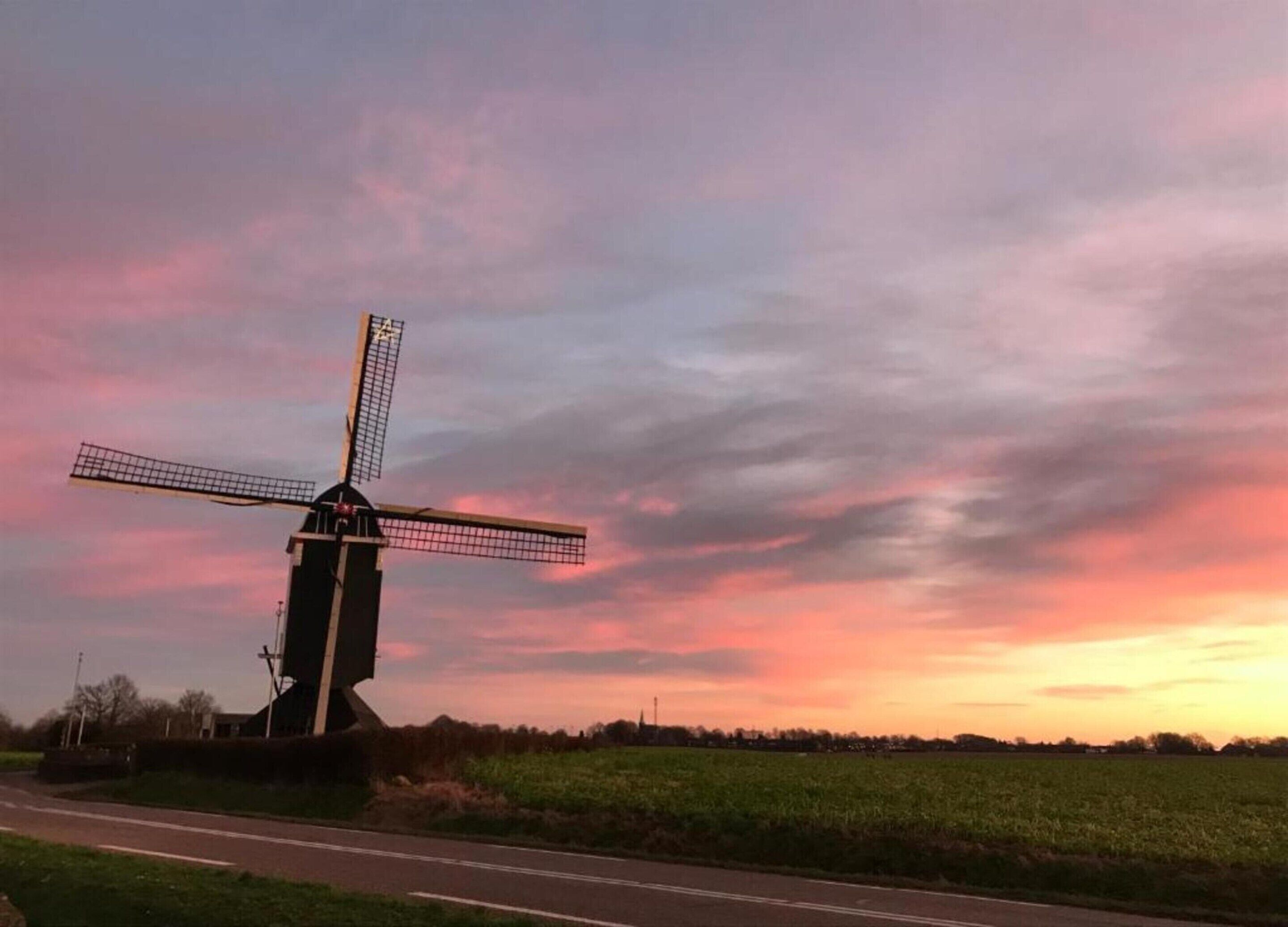 A windmill in a rural setting with a road in the foreground and a sunset in the background.
