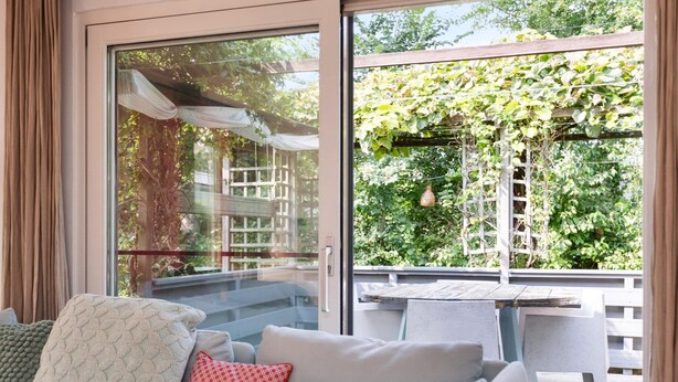 Sala moderna com sofá, almofadas estampadas e vista para um jardim através de grandes portas de vidro.