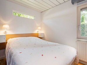 A bedroom with a wooden headboard, two bedside lamps, a window with a view of greenery, and a bed with a white bedspread featuring small colorful flowers.