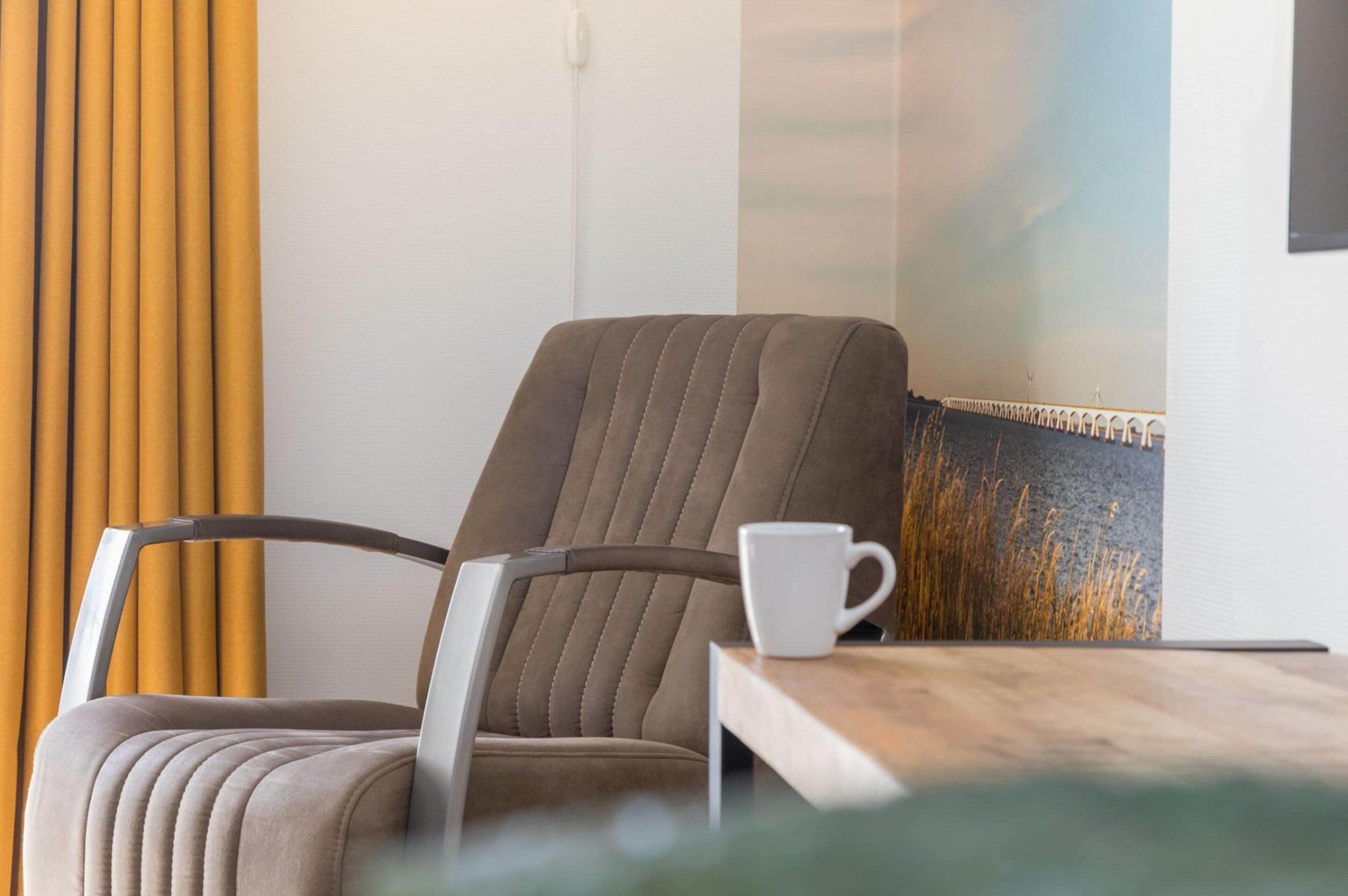 Una silla con respaldo y reposabrazos acolchados, una taza blanca sobre una mesa de madera, y una cortina amarilla al fondo.