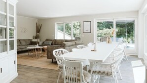 A spacious living room with a large white dining table, grey sofa, and white chairs. There is a glass cabinet on the left and a large window providing a view of greenery outside. - Luxurious 16-person Villa With Swimming Pool Sauna Outdoor Kitchen With BBQ (Ouddorp)