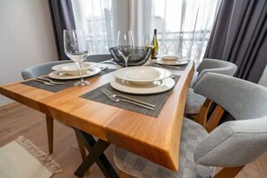A dining table set with plates, cutlery, and wine glasses, with a view of the city through large windows. - Pomorie Seaside Apartments (Pomorie)