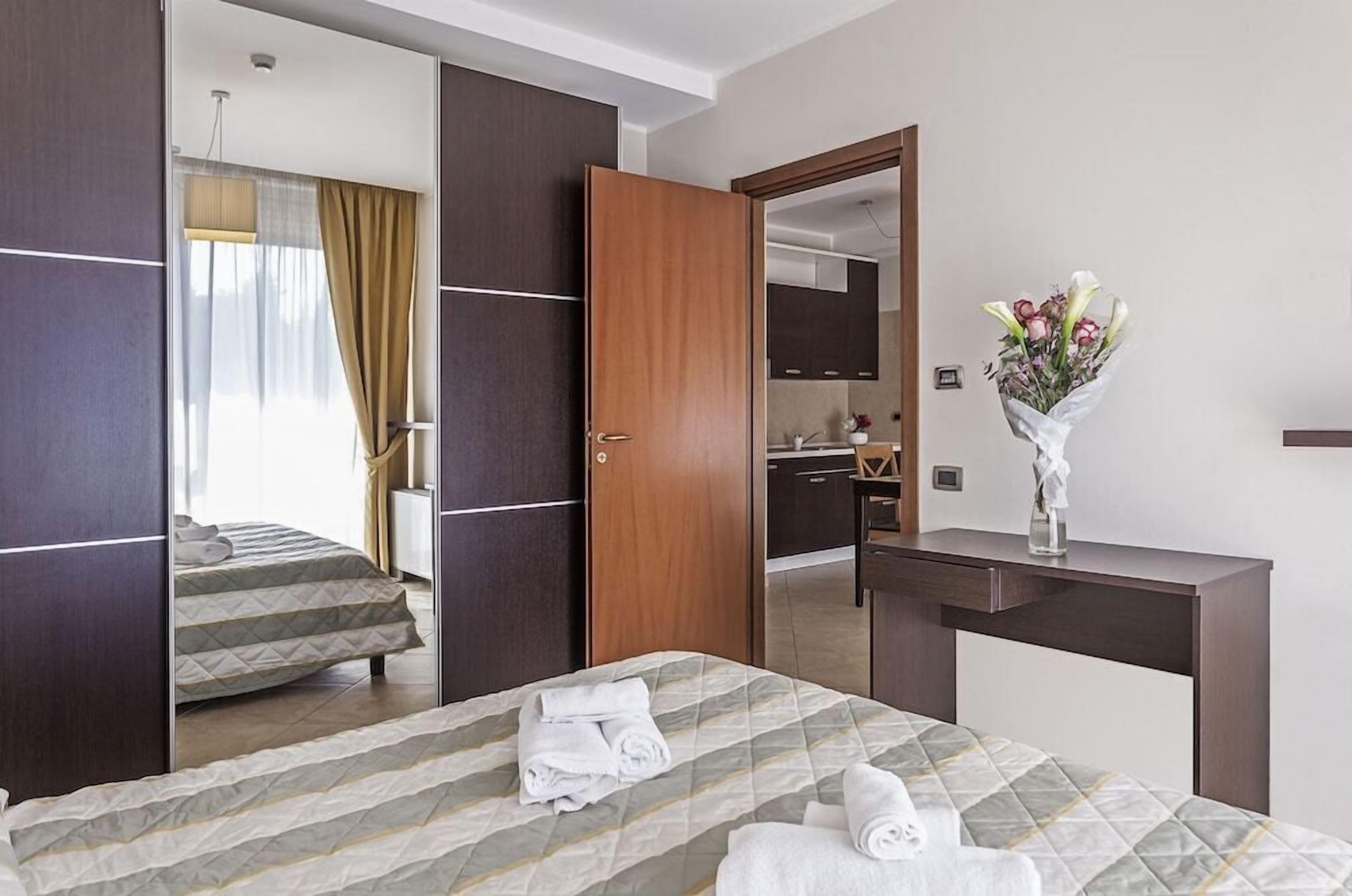A hotel room with a bed, a desk, a mirror, and a bouquet of flowers.