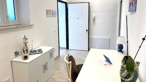 A room with a white desk, a chair, a small cabinet with decorative items, a door, and a window. - Villa in Sicily With Sea Access (Santa Flavia)