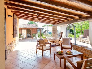 A covered patio area with wooden furniture, a stone wall, and a view of a house with a tiled roof.