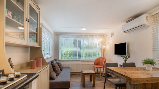 A compact kitchen with a built-in stove, a dining area with a wooden table and chairs, a sofa, and a large window with blinds. - Beautiful Holiday Chalet in Emst With a Private Garden (Emst)