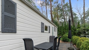 A white wooden house with black shutters, a patio with a table and chairs, and a garden with trees and shrubs. - Beautiful Holiday Chalet in Emst With a Private Garden (Emst)