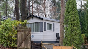 A small, single-story house with a white exterior and dark shutters, surrounded by dense trees and a wooden gate. - Beautiful Holiday Chalet in Emst With a Private Garden (Emst)