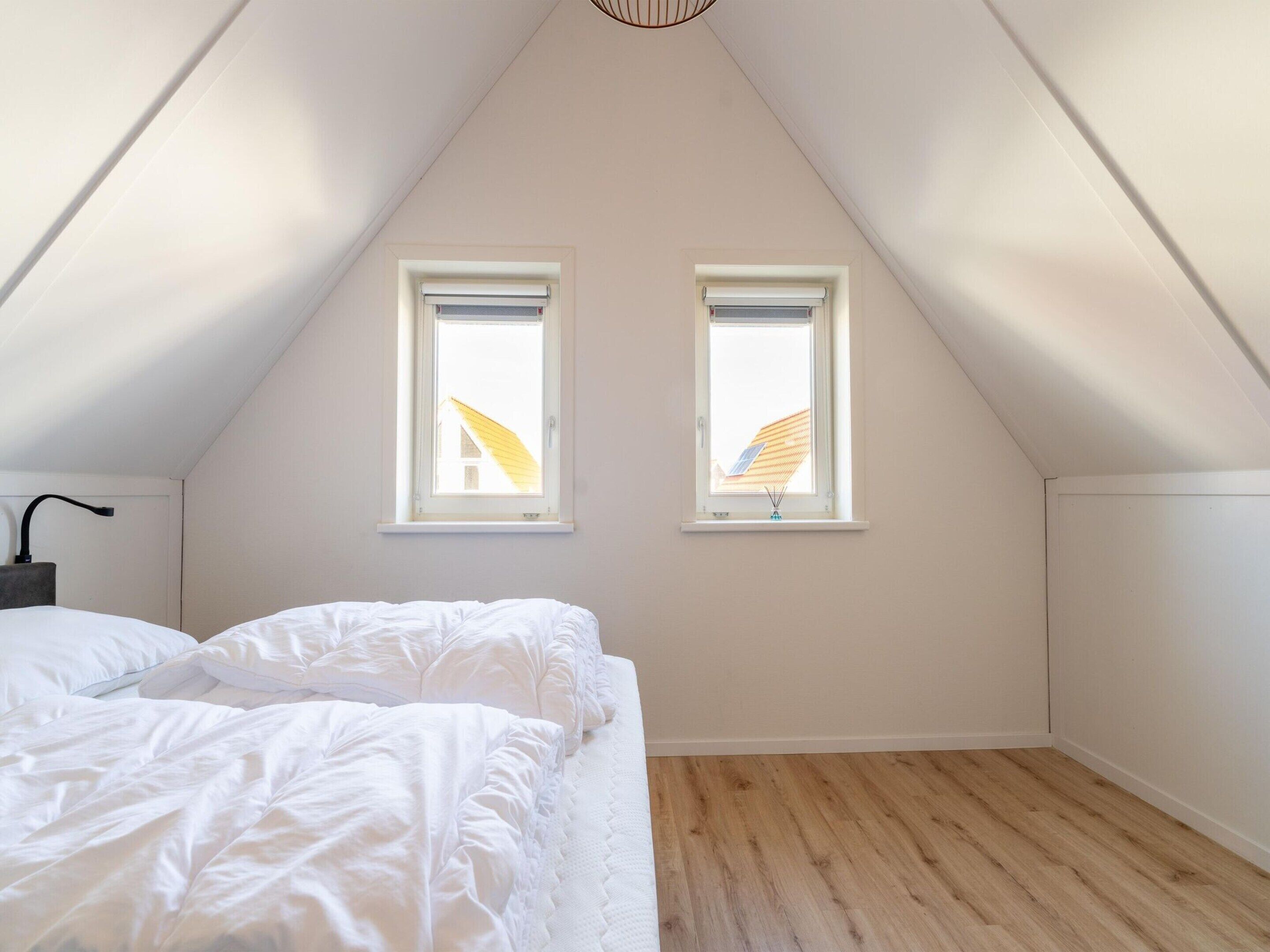 A bedroom with a sloped ceiling, two windows, a bed with white bedding, and a wooden floor.
