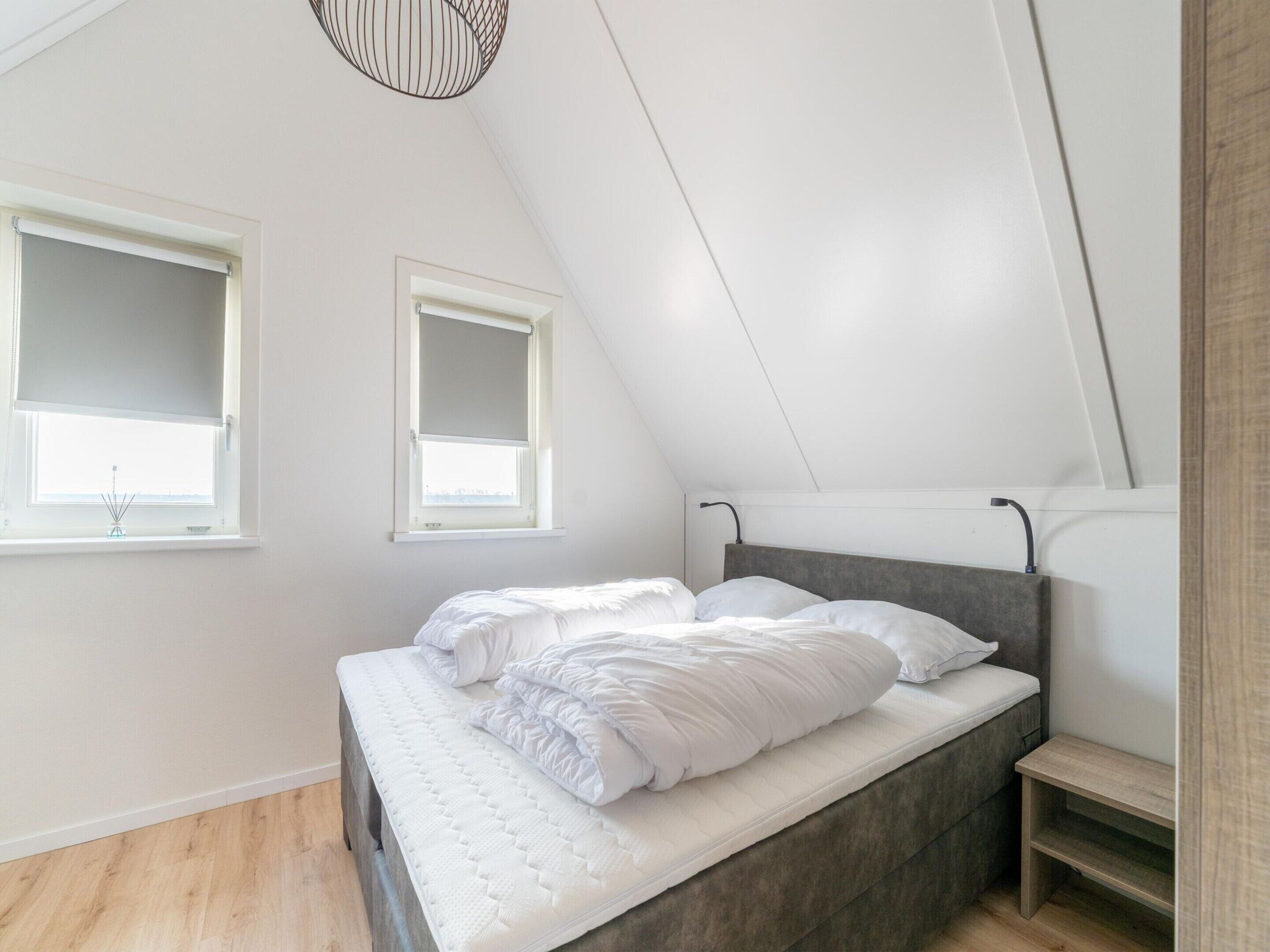 A modern bedroom with a sloped ceiling, a bed with white bedding, two windows with blinds, and a small wooden shelf.