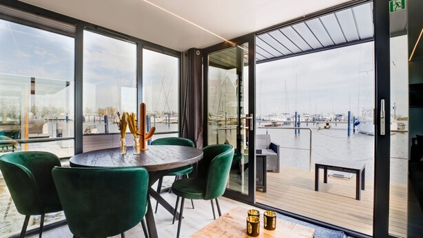 Ein moderner Essbereich mit einem runden Tisch, grünen Stühlen und Blick auf einen Yachthafen durch große Fenster.