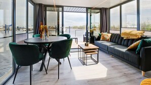 Ein modernes Wohnzimmer mit einem runden Tisch, grünen Sesseln, einer Couch und großen Fenstern mit Blick auf das Wasser.
