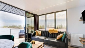 Ein modernes Wohnzimmer mit einer Couch, einem Tisch, Stühlen und großen Fenstern mit Blick auf das Wasser. Ein Fernseher ist an der Wand befestigt.