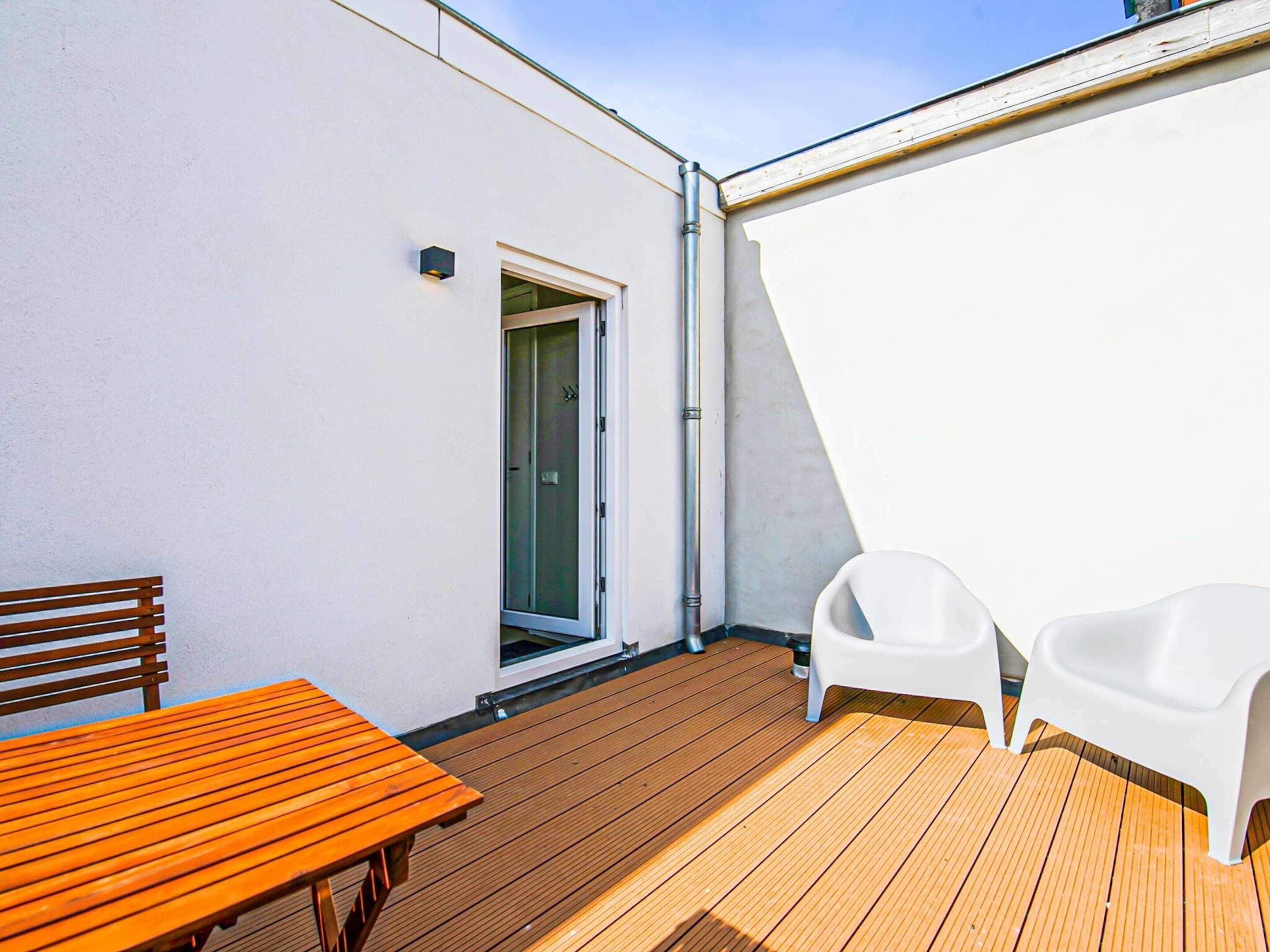 A wooden deck with a table and two white chairs, a sliding glass door, and a white wall.