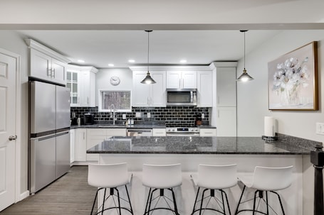 A modern kitchen with white cabinets, a stainless steel refrigerator, a black countertop, and three white bar stools.