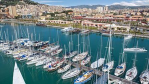A marina with numerous sailboats docked, a cityscape in the background, and a prominent hill with a castle-like structure. - Belvilla by OYO Acanto Bilocale (Modica)