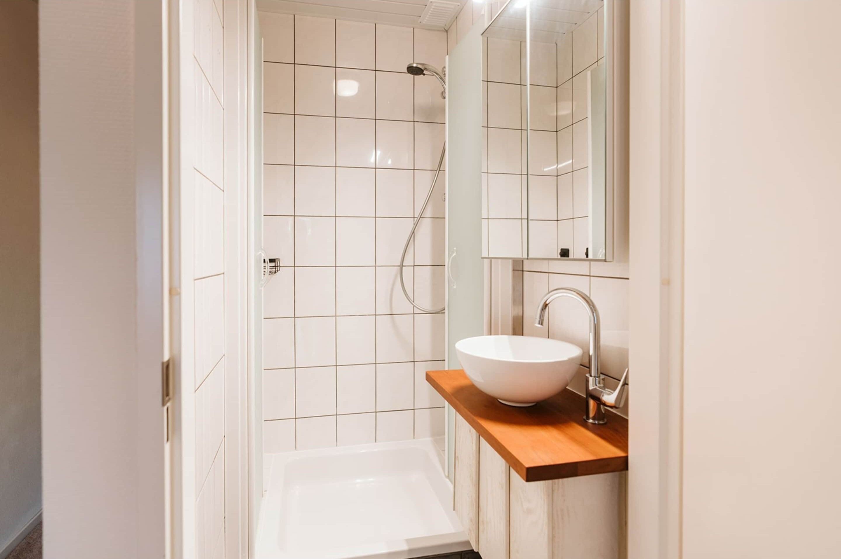 A modern bathroom with a white tiled shower, a white sink with a wooden countertop, and a mirror above the sink.