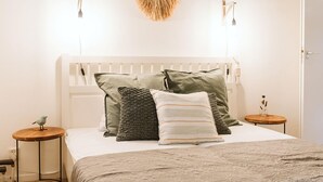 A neatly made bed with a white headboard, multiple pillows, and a decorative wall hanging. There are two bedside tables with lamps and a small round table with a decorative item.