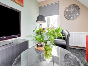 A modern living room with a flat-screen TV, a round glass table with a vase of flowers, a black chair, and a wall clock. - Peaceful Family Home in Elim (Elim)