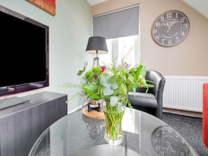 A modern living room with a flat-screen TV, a round glass table with a vase of flowers, a black chair, and a wall clock. - Peaceful Family Home in Elim (Elim)