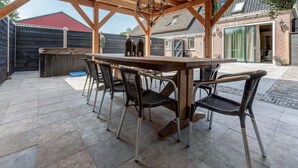 An outdoor patio with a wooden table and chairs, a hanging light fixture, and a view of a brick house in the background. - Stylish Farmhouse With Spa Tub (Kootstertille)