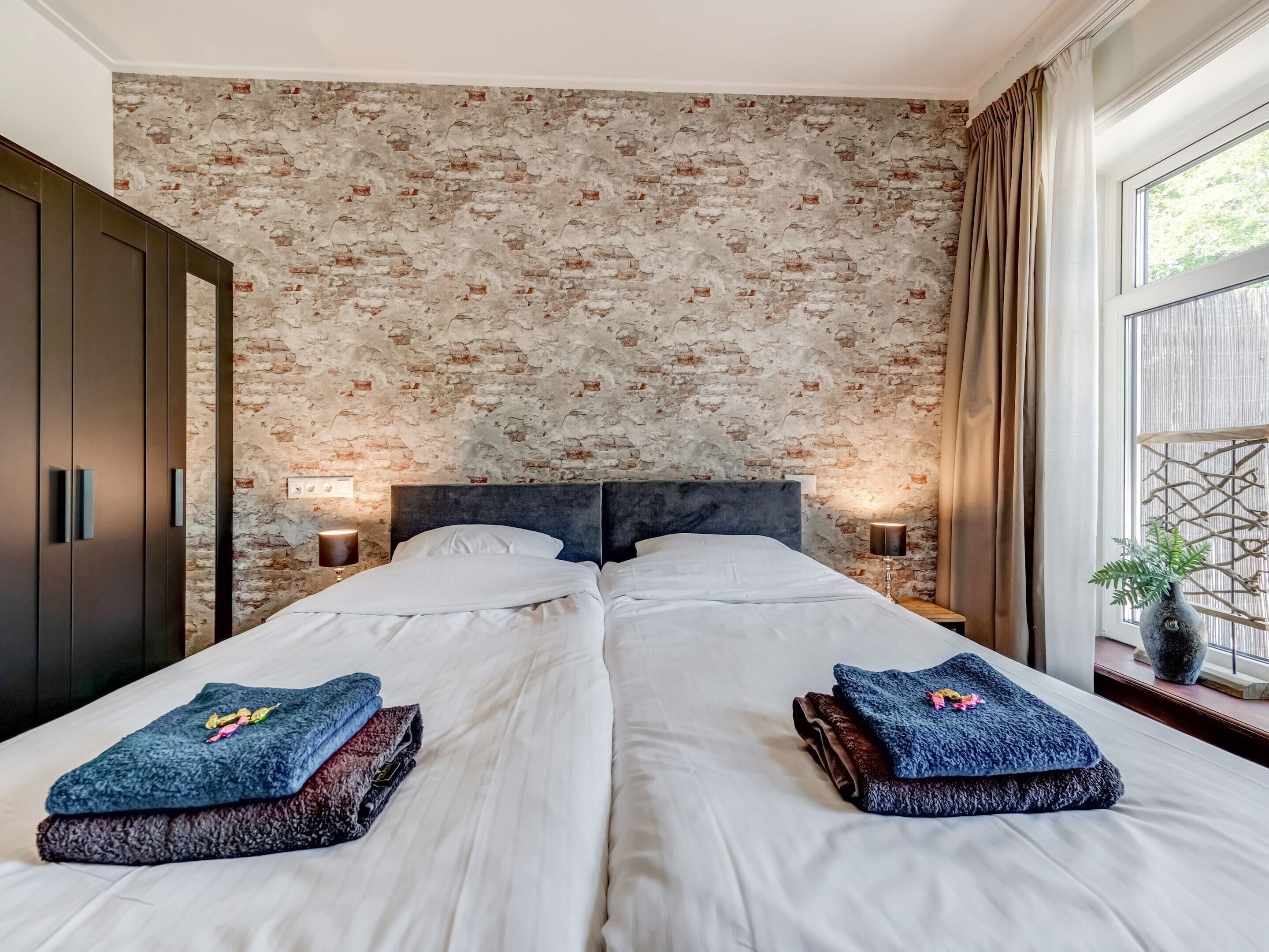 A double bed with white linens, two folded towels, a brick-patterned wall, a dark headboard, bedside lamps, and a window with curtains.