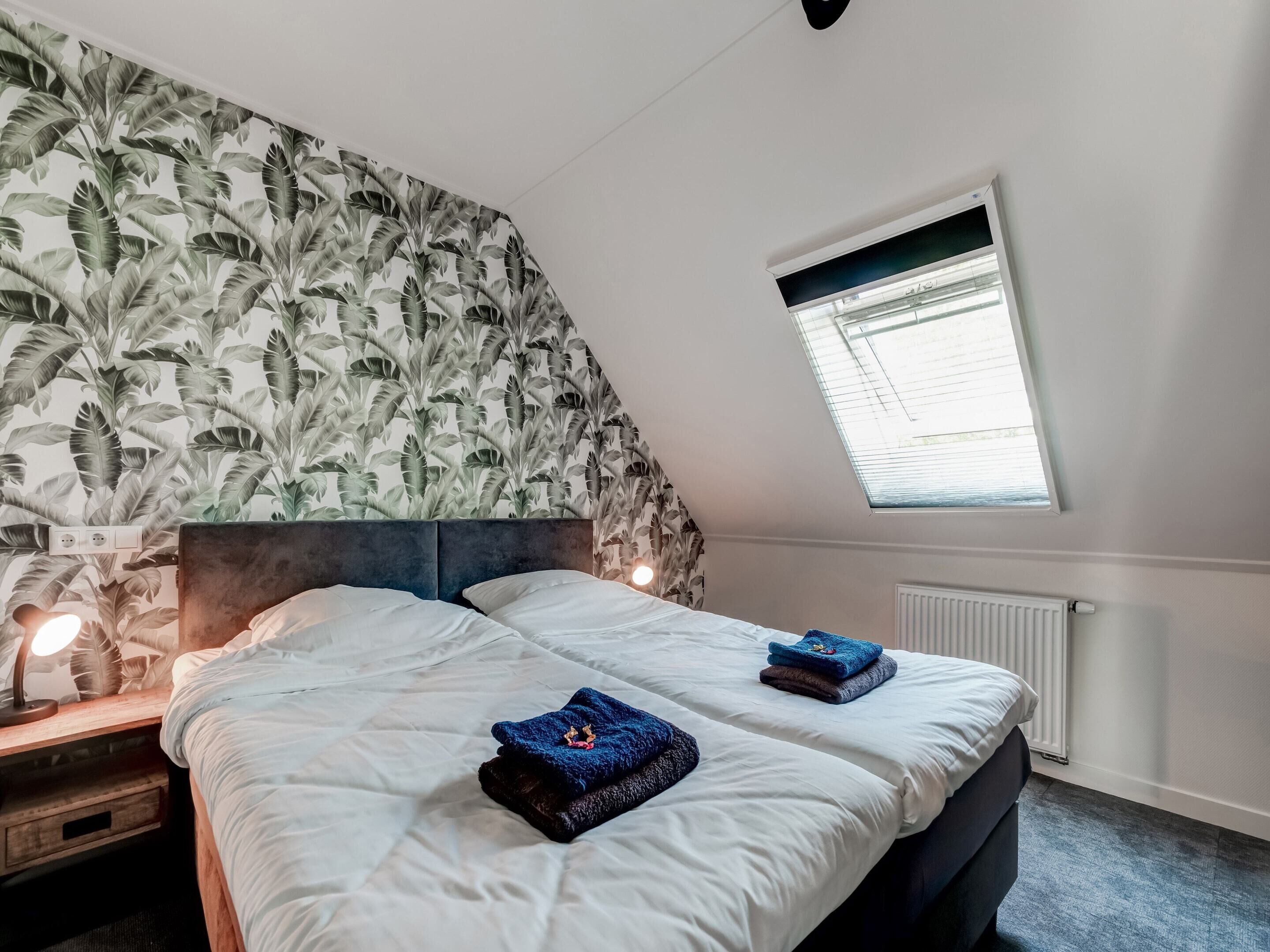 A bedroom with two beds, a sloped ceiling, a window with blinds, a radiator, and a wall with a leaf pattern.