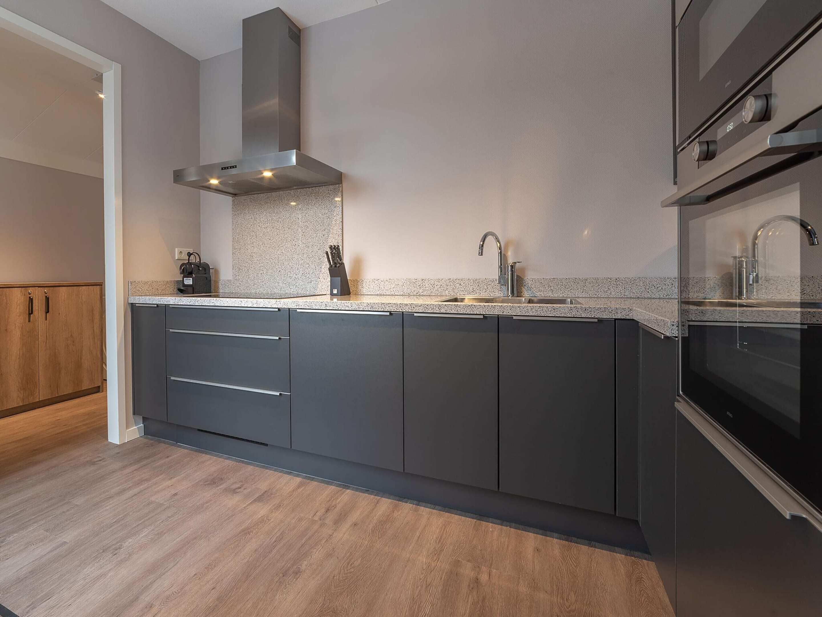 A modern kitchen with dark cabinetry, a stainless steel range hood, a granite countertop, and a built-in oven.
