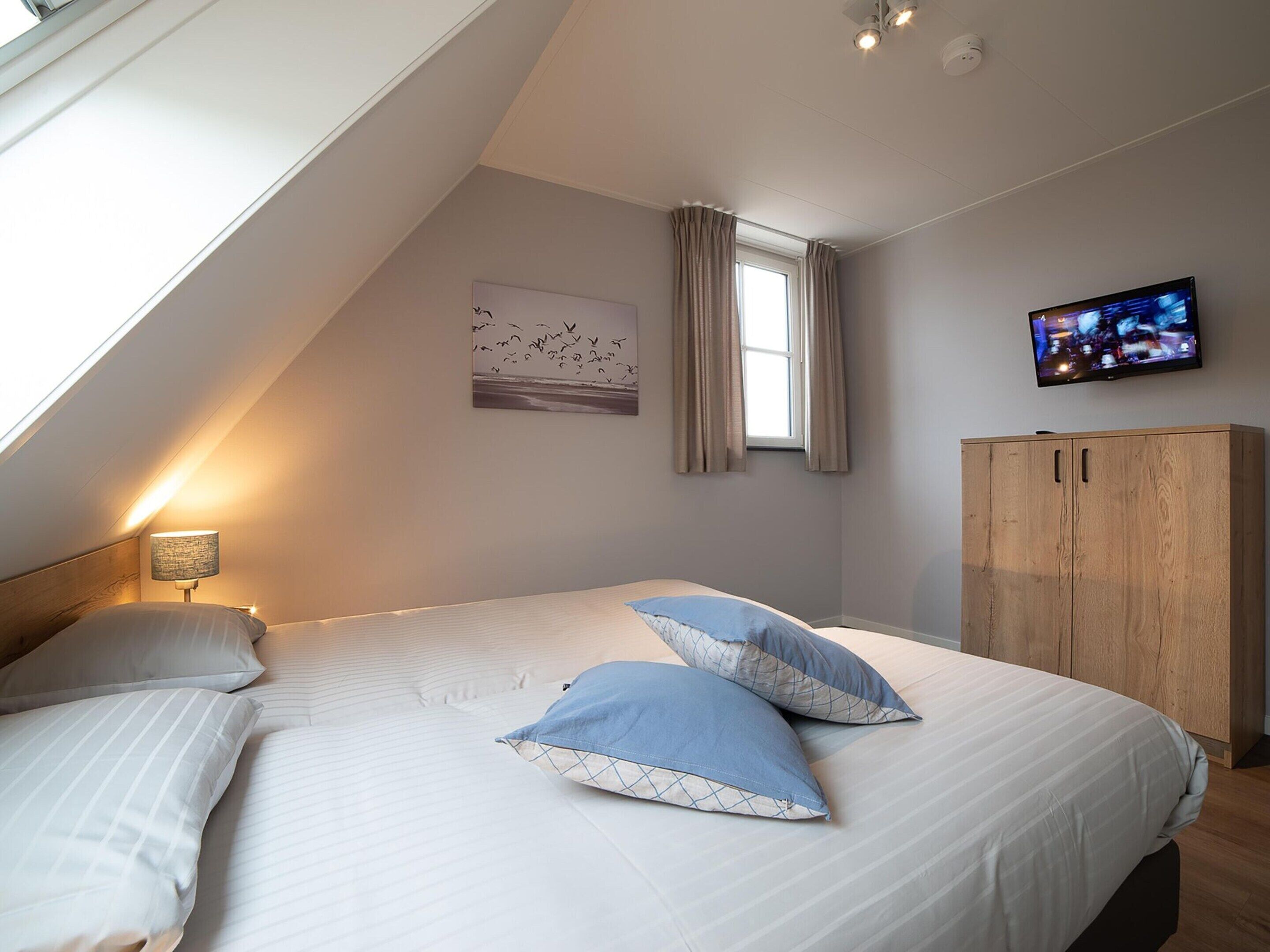 A bedroom with a sloped ceiling, a bed with two pillows, a wooden wardrobe, a television on the wall, and a window with curtains.