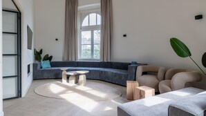 Een moderne woonkamer met een gebogen grijze bank, een ronde salontafel en een beige fauteuil. Er is een groot raam met gordijnen, een potplant en een uitzicht op gebouwen.