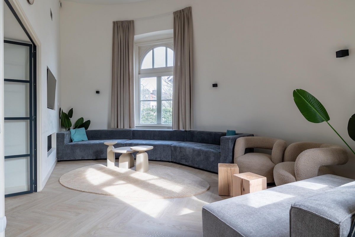 Un salon moderne avec un canapé gris aux lignes courbes, une table basse ronde et un fauteuil beige. Une grande fenêtre, ornée de rideaux, laisse entrer la lumière, et on aperçoit une plante en pot ainsi qu’une vue sur les bâtiments environnants.