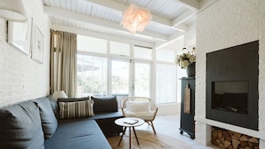A modern living room with a dark grey sofa, a white armchair, a round coffee table, a fireplace, and large windows with curtains. - Lovely Holiday Home 10 Minutes by Bike (Ouddorp)